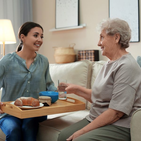 A young woman in a striped blouse brings a breakfast tray to a smiling older woman on a sofa at home